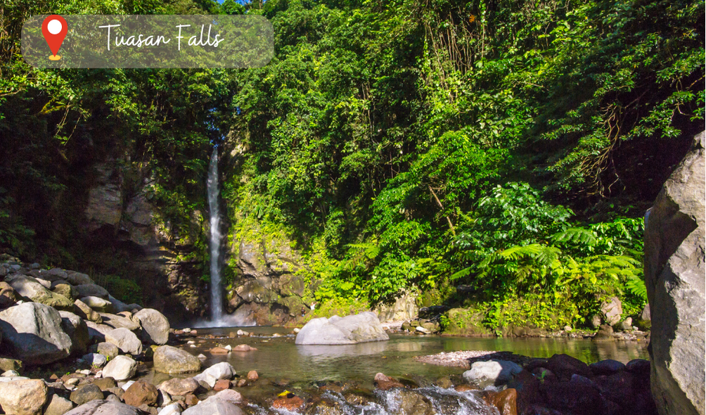 The image captures Tuasan Falls, a stunning waterfall in Camiguin Island, Philippines. The water cascades down a series of rocky steps, surrounded by lush green foliage and towering trees. The pool at the base of the falls is crystal clear and inviting, with people seen swimming and lounging around. The sunlight filters through the trees, casting a gentle glow over the entire scene. It's a serene and picturesque view that showcases the natural beauty of the Philippines. #TuasanFalls #CamiguinIsland #Philippines #Waterfall #Nature #Landscape #Swimming #Relaxation #TravelPH